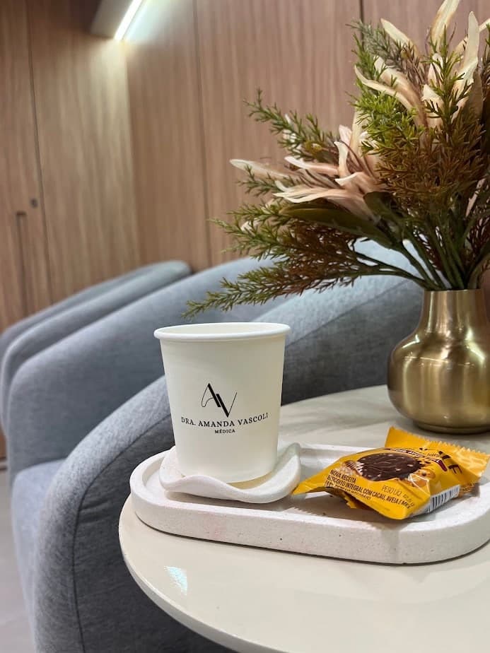 Detalhe da sala de espera: café servido em copo com a marca da clínica, ao lado de um arranjo de flores
